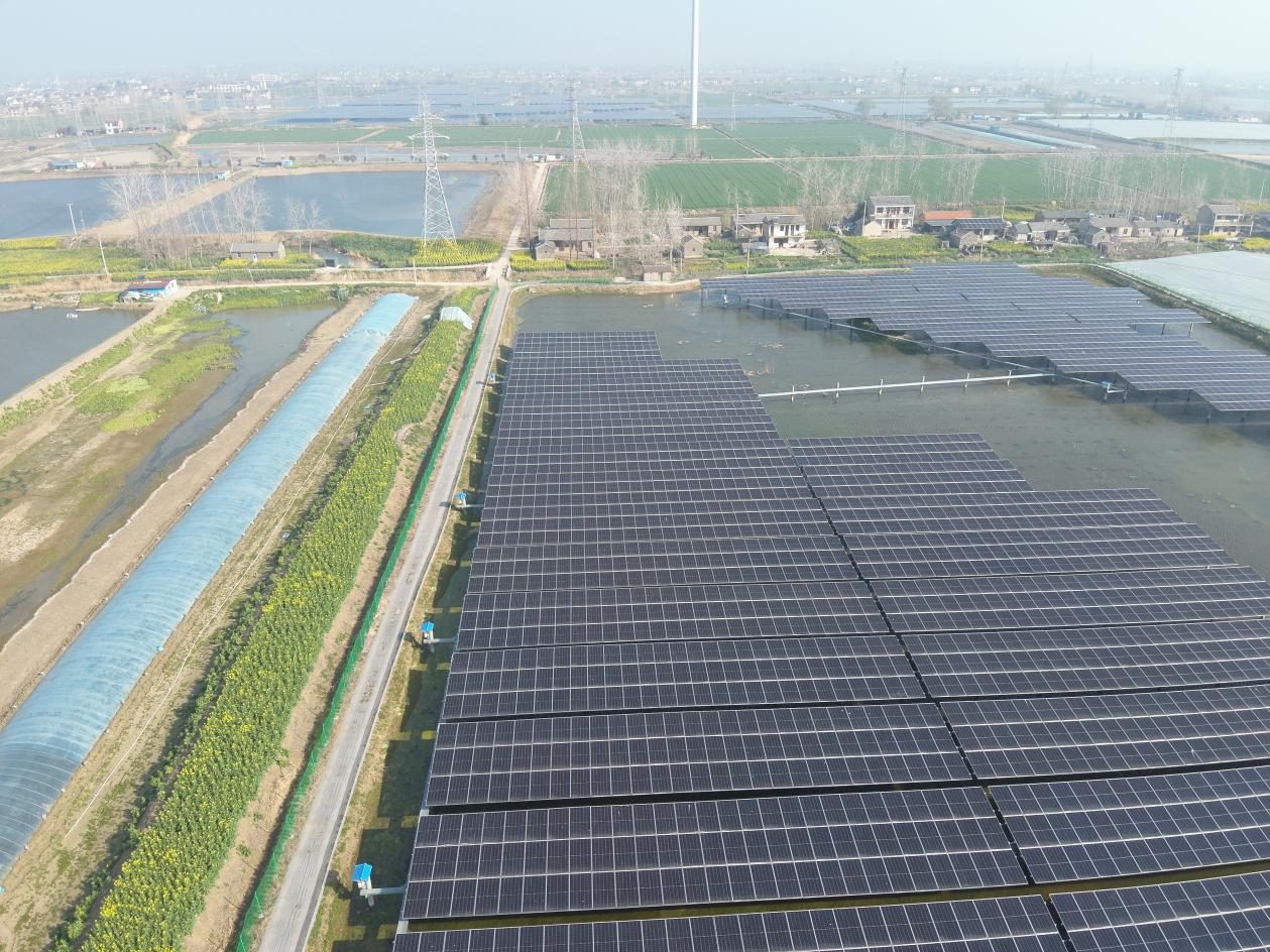 Aerial view of a solar power plant beside farmland, ponds and a greenhouse, with a wind turbine in the distance